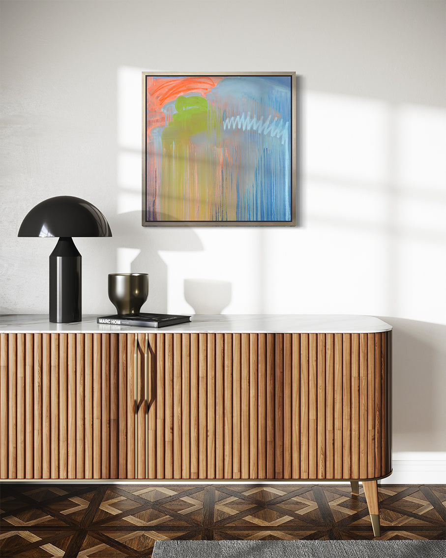 A blue, orange and green abstract print framed in a white oak floater frame by Teodora Guererra hanging on a white wall over a mid century modern console with a black lamp.