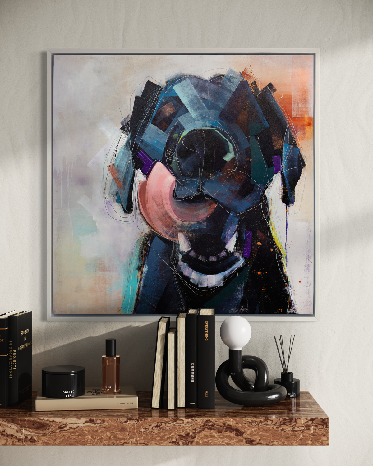 A dark blue, black, purple and copper abstract dog print in a white floater frame hangs on a white wall above a copper granite floating shelf with stacks of books and decorative accessories.