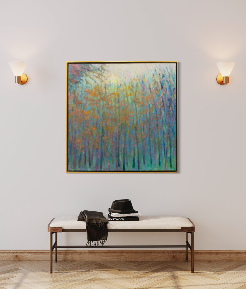 A colorful abstracted landscape painting framed in a gold frame hangs on a white wall between two gold wall sconces and above a modern wood bench with a white cushion, on which rests a scarf, book, and hat. 