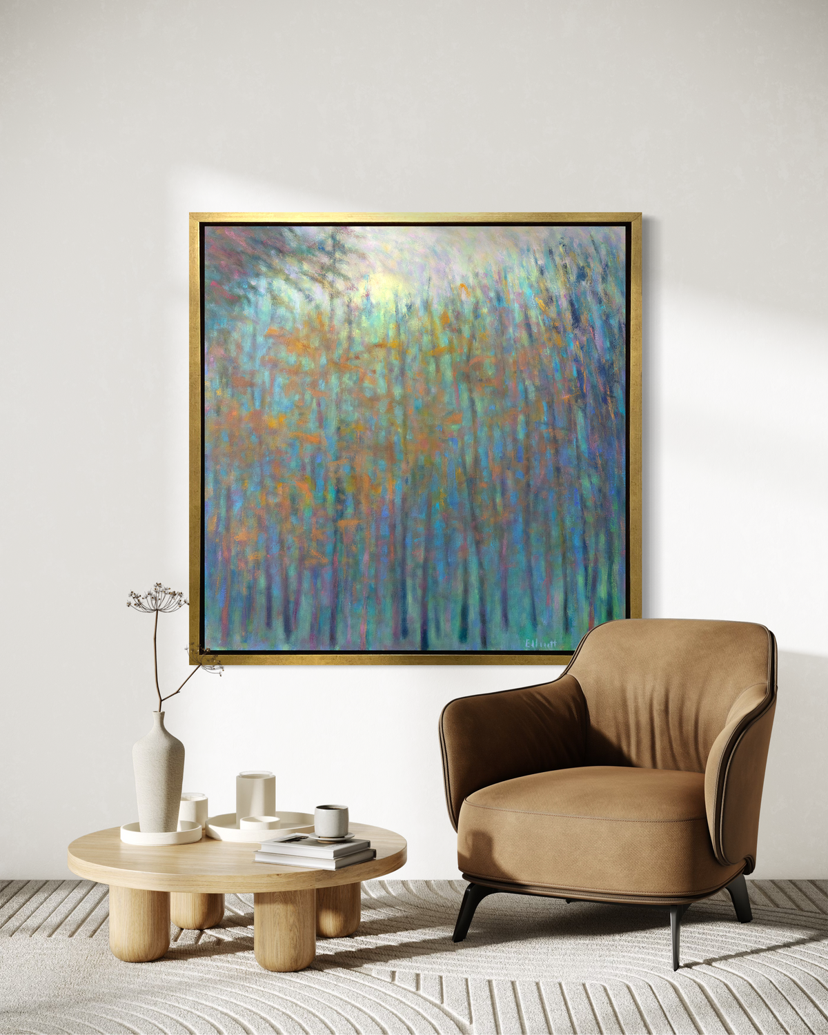An impressionist landscape print in a gold floater frame hangs on a white wall above a soft brown armchair and a low round coffee table with decorative accessories.