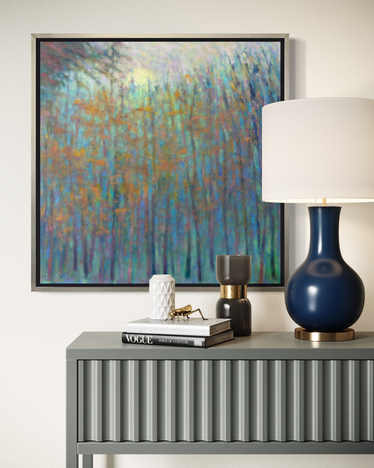 An impressionist landscape print in a silver floater frame hangs on a white wall above a grey console table with a blue table lamp and a stack of books.