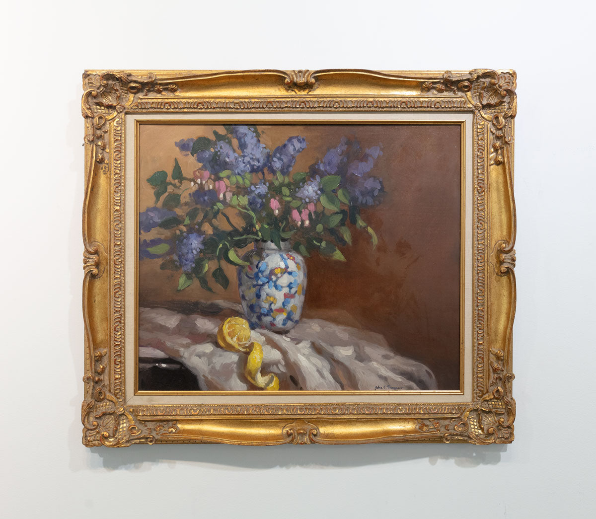 Hyperrealist painting of lilacs in a vase with peeled lemon underneath. Painting displayed in gold frame on white wall.