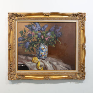 Hyperrealist painting of lilacs in a vase with peeled lemon underneath. Painting displayed in gold frame on white wall.