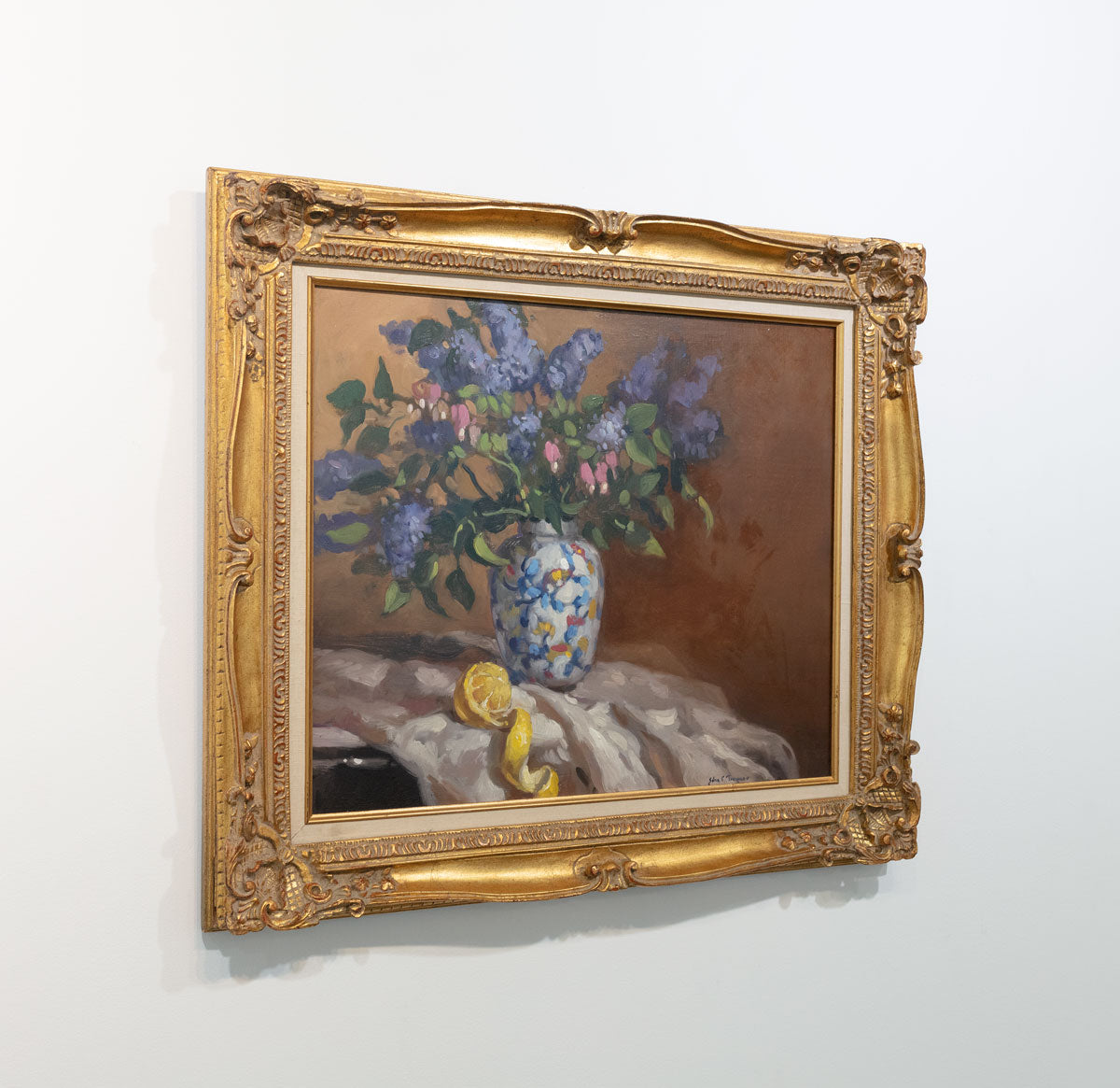 Hyperrealist painting of lilacs in a vase with peeled lemon underneath. Painting displayed in gold frame on white wall viewed from the left.