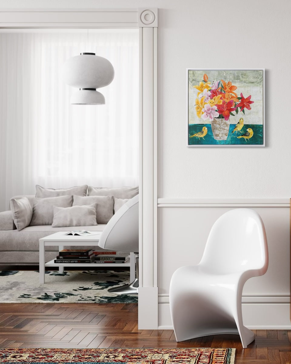 A framed square collage painting of a colorful floral bouquet hangs on a white wall in a modern living room setting with a white chair, sofa, and wood flooring.