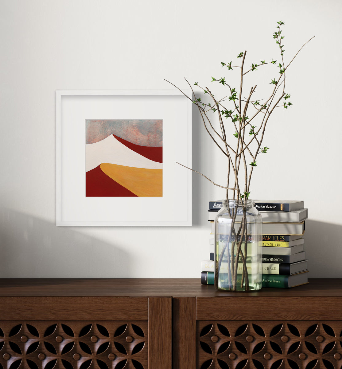 A dark red, white, dark yellow and teal abstract landscape print in a white frame with a mat hangs on a white wall above a dark wood credenza with a stack of books and a clear jar with greenery stems.