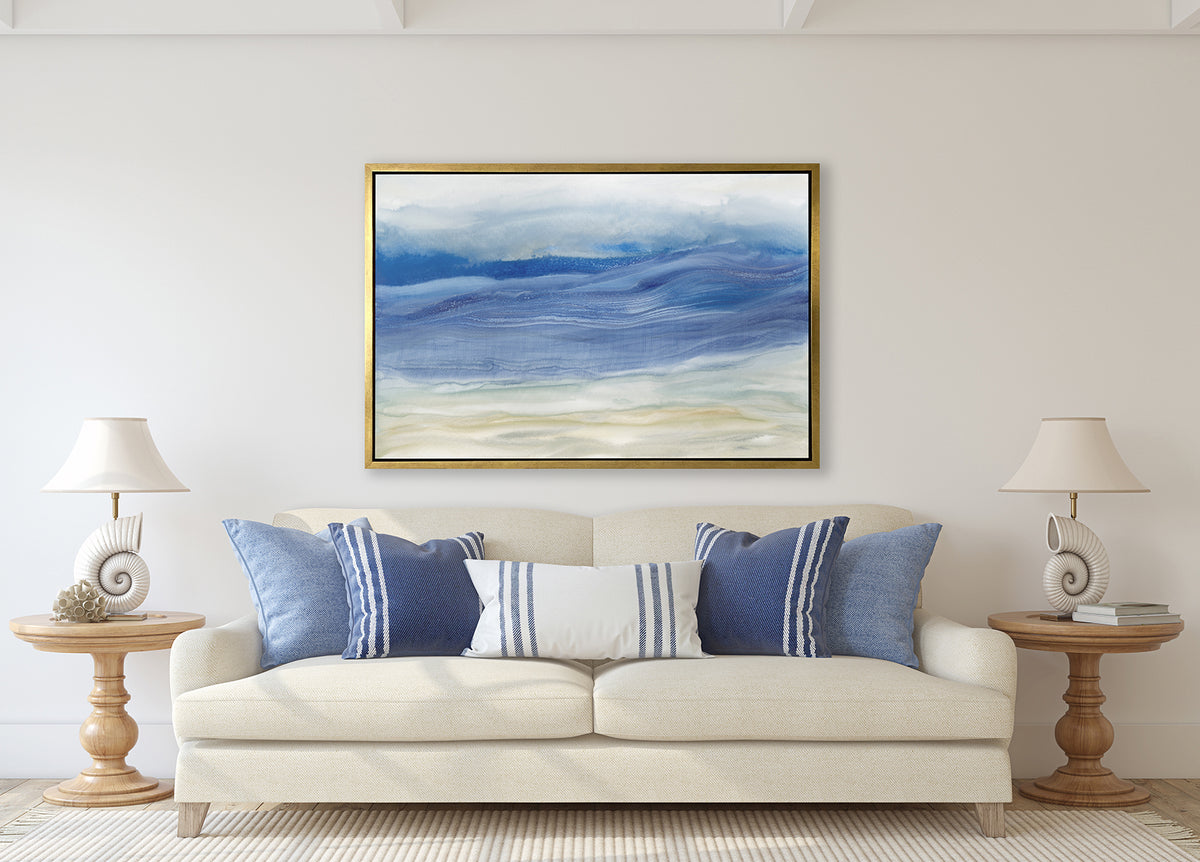 A blue, white and beige abstract print in a silver floater frame hangs on a gold wall above a cream linen sofa with blue and white pillows and two matching wood pedestal side tables with matching white seashell table lamps.