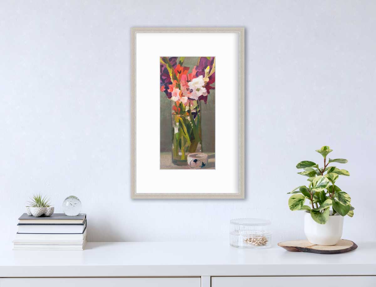 A purple, pink and green abstract floral print in a silver frame with a mat hangs on a white wall above a white surface with a stack of books, a clear round container with gold accessories and a potted plant.