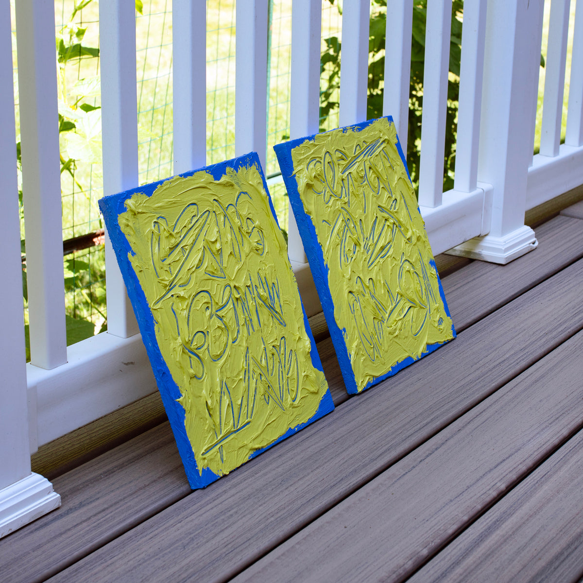 Two abstract text based painting featuring a thick, yellow-green impasto texture with blue abstract scribbled lines and a bright blue background. They are displayed on a wooden deck, side view, in front of a white railing with green foliage behind.