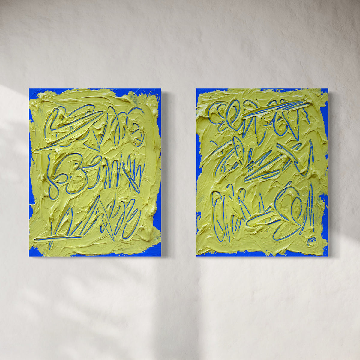 Two abstract text based paintings hanging side by side on a textured white wall. Each canvas features a thick, yellow-green impasto texture with blue abstract scribbled lines and a bright blue border.