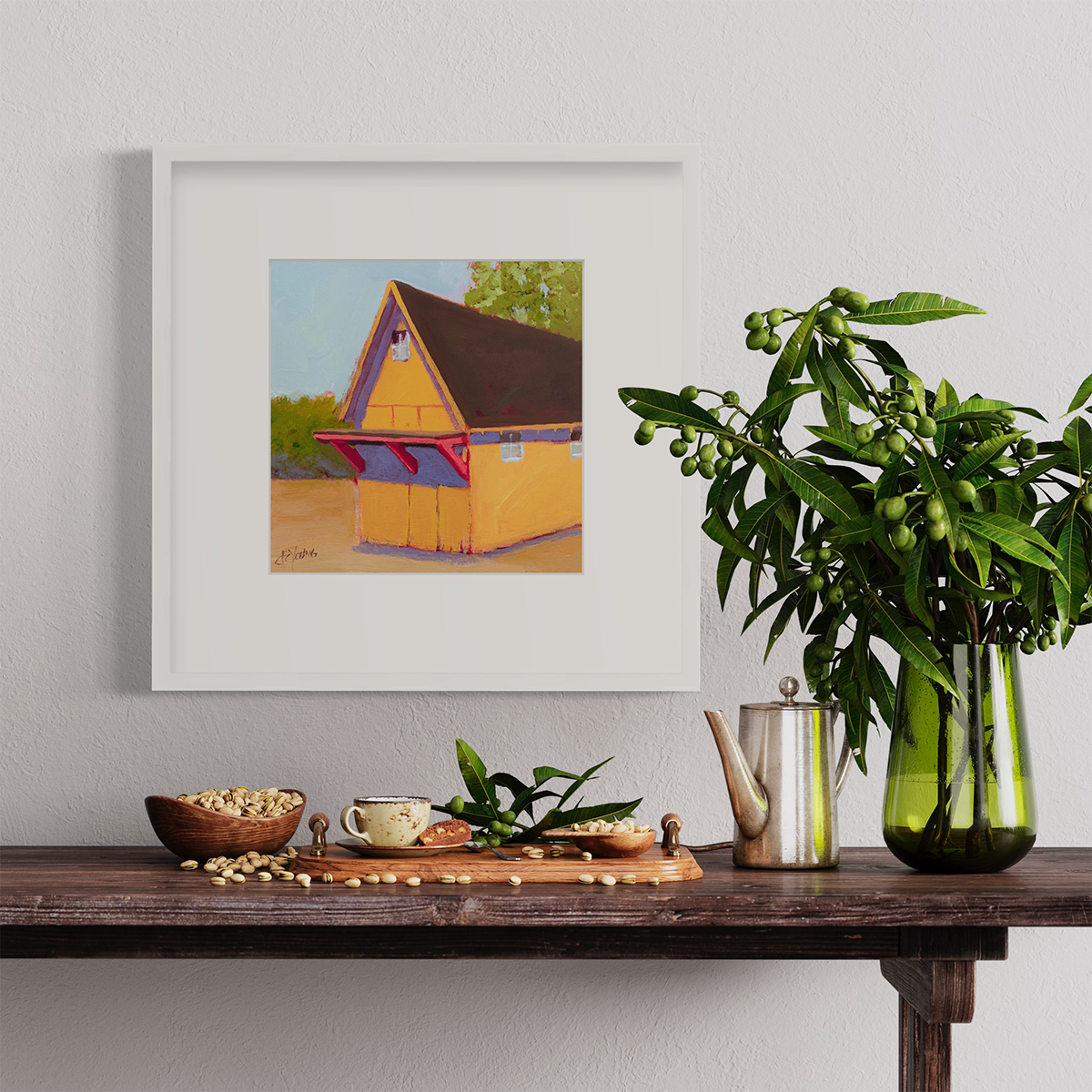 A yellow, orange, lavender and blue contemporary barn print in a white frame with a mat hangs on a white wall above a wood shelf with a tray of pistachios, a small metal kettle and a green glass vase with greenery. 