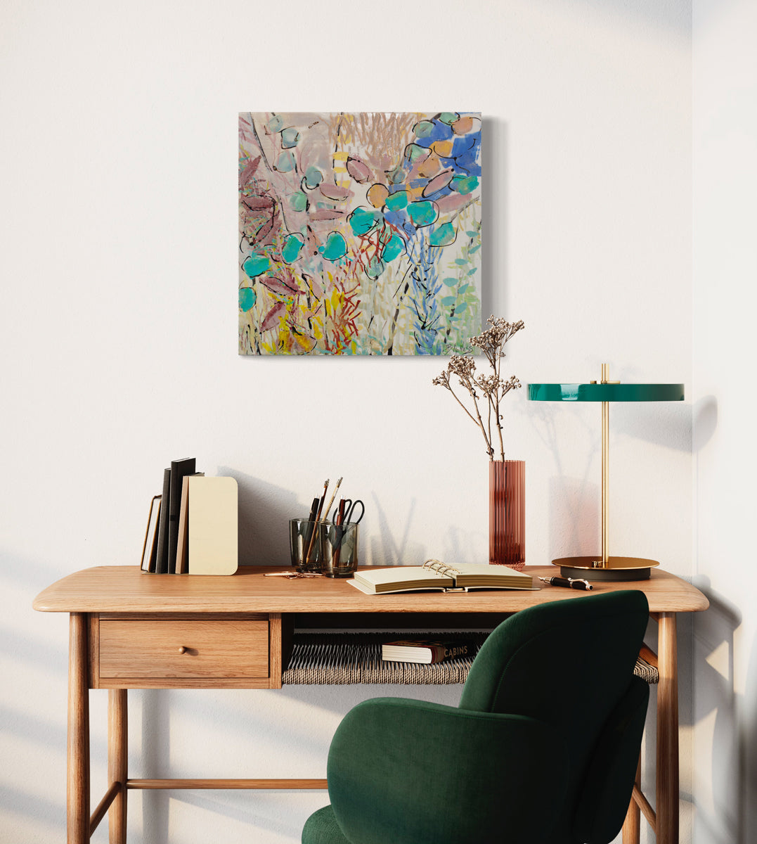 A colorful pink, yellow, and turquoise abstract painting hangs on a white wall above a wooden desk with table decor, an open notebook, and green table lamp in front of a green desk chair. 