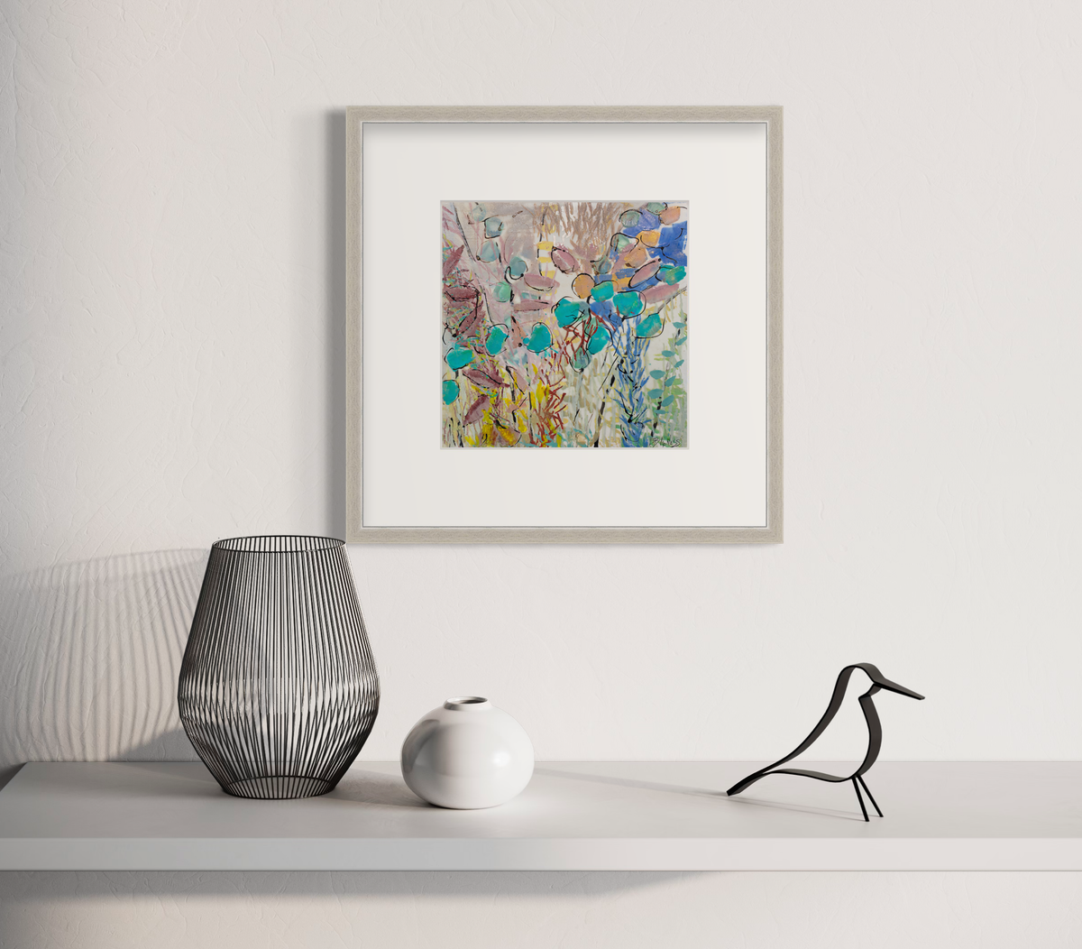 A blue, pink and yellow abstract floral print in a silver frame with a mat hangs on a white wall a white shelf with a black wire vase, a small white vase and a bird decoration.