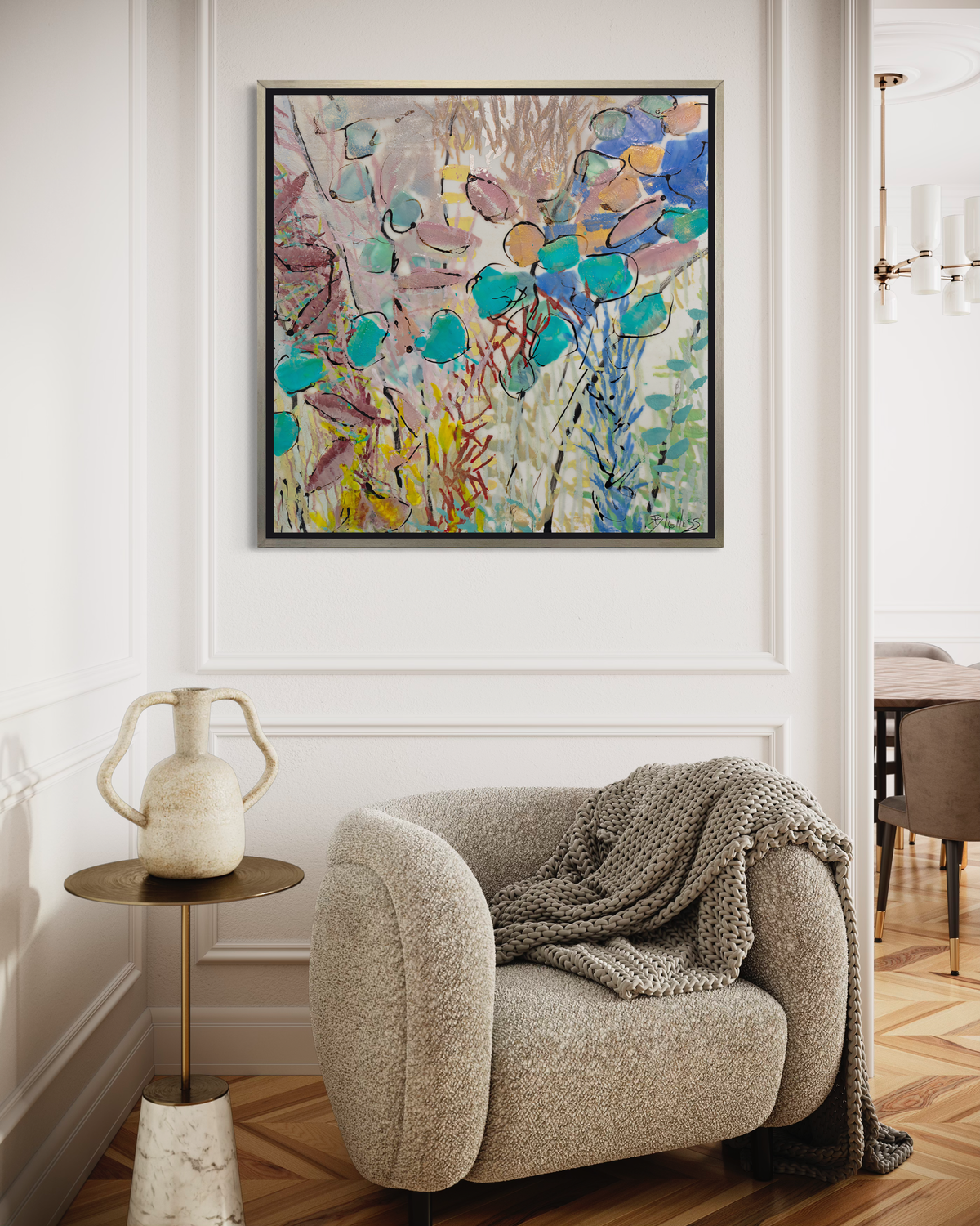 A blue, pink and yellow abstract floral print in a silver floater frame hangs on a white paneled wall above a low modern cream armchair with a throw blanket and a side table with a decorative jar.