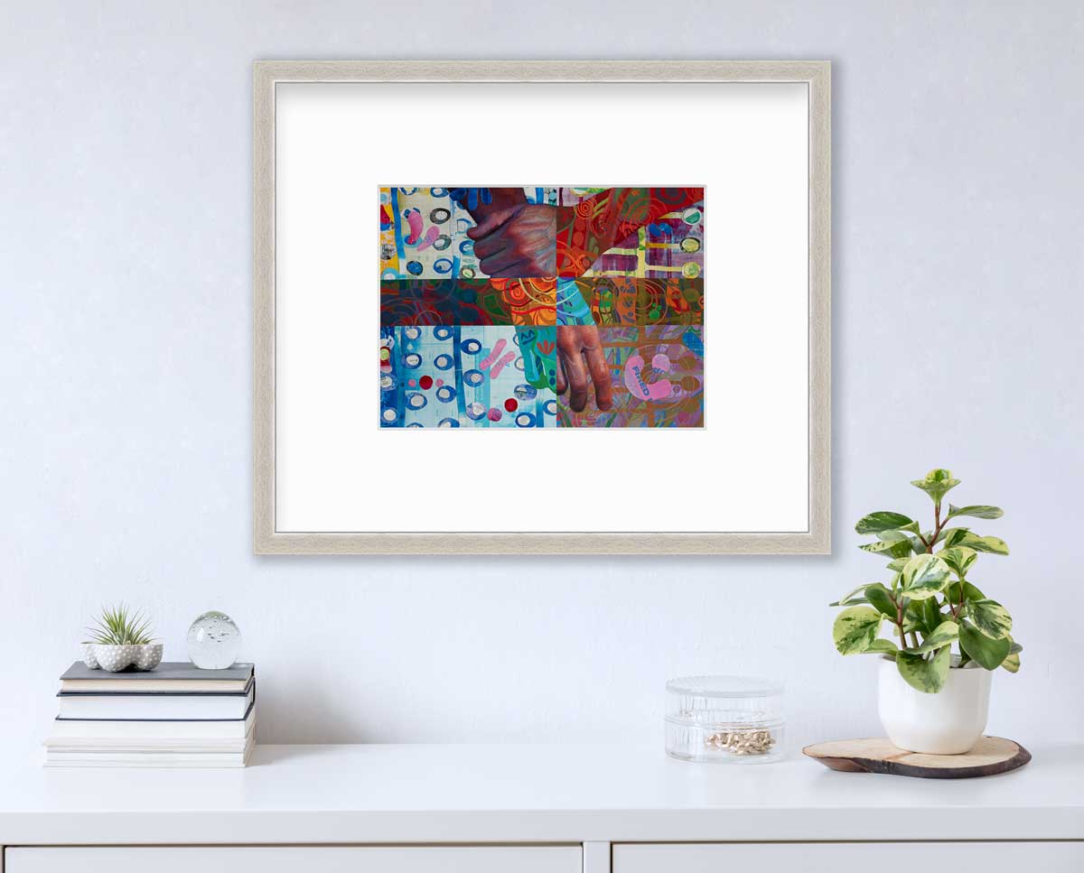 A multicolored abstract print of two hands in a silver frame with a mat hangs on a white wall above a stack of books, a clear round container with gold accessories and a potted plant.