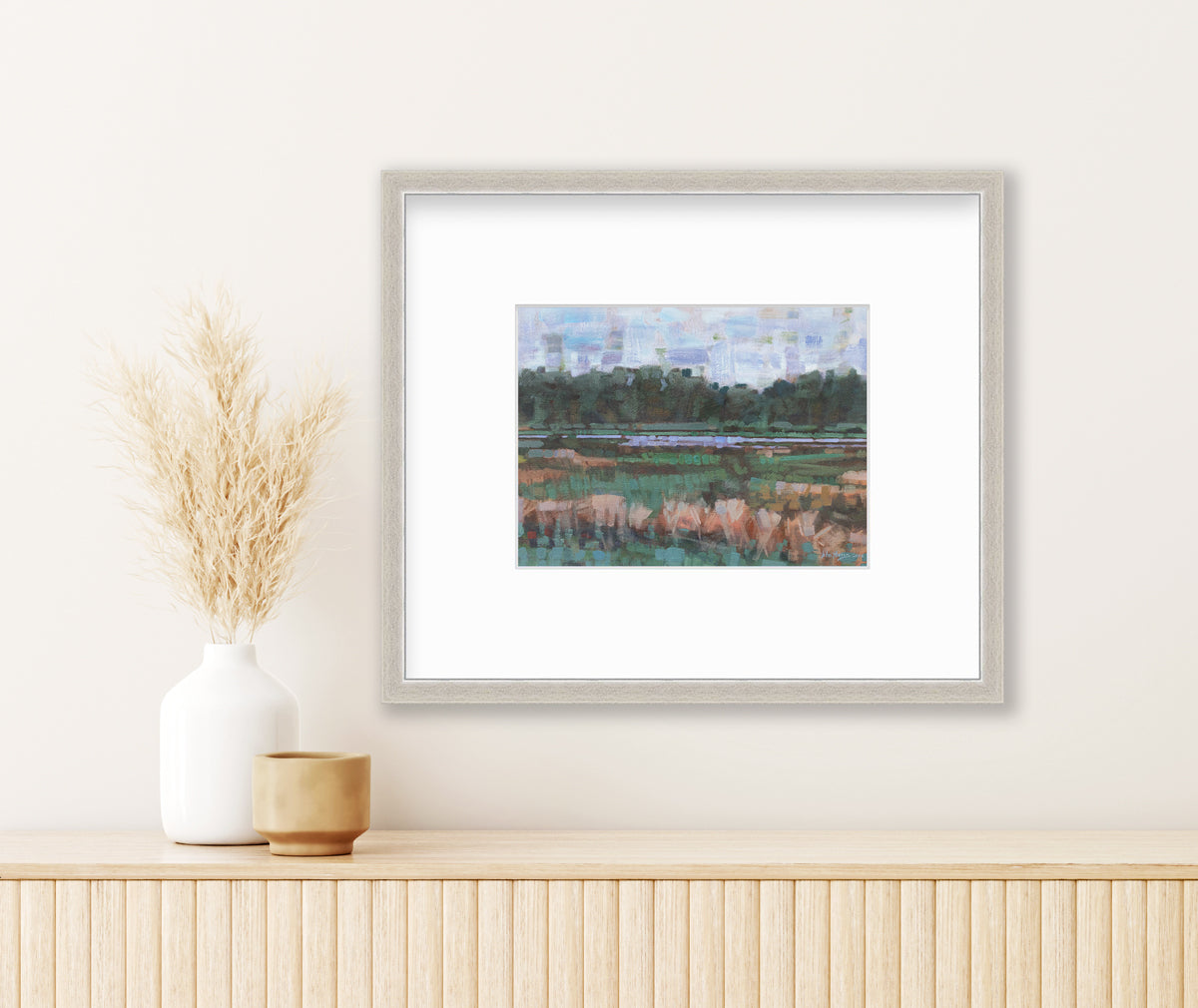 A green, orange and blue abstract landscape print in a silver frame with a mat hangs on a white wall above a light wood surface with a beige decorative dish and a white vase with wheat stems.