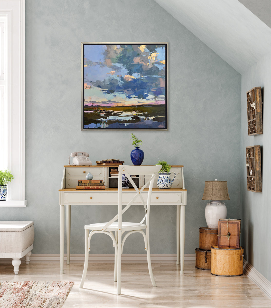 A blue coastal landscape art print framed in a silver frame hangs on a grey wall above a traditional white desk with vintage decor. 