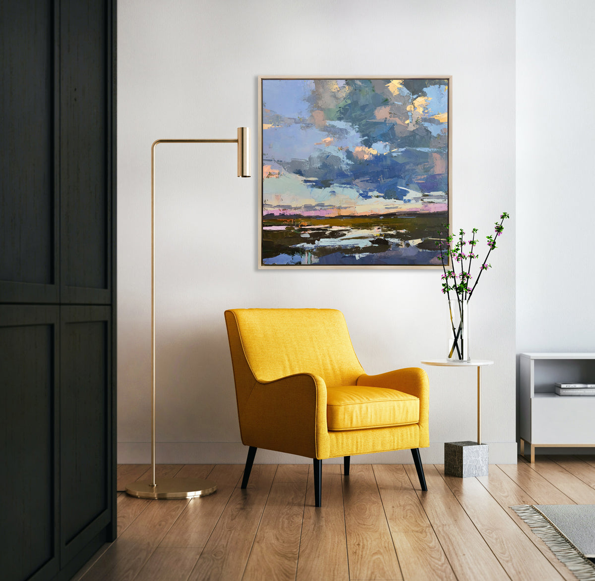 A blue coastal landscape art print framed in a white oak frame hangs on a white wall above a modern yellow armchair, floor lamp, and side table with a vase with flowers. 