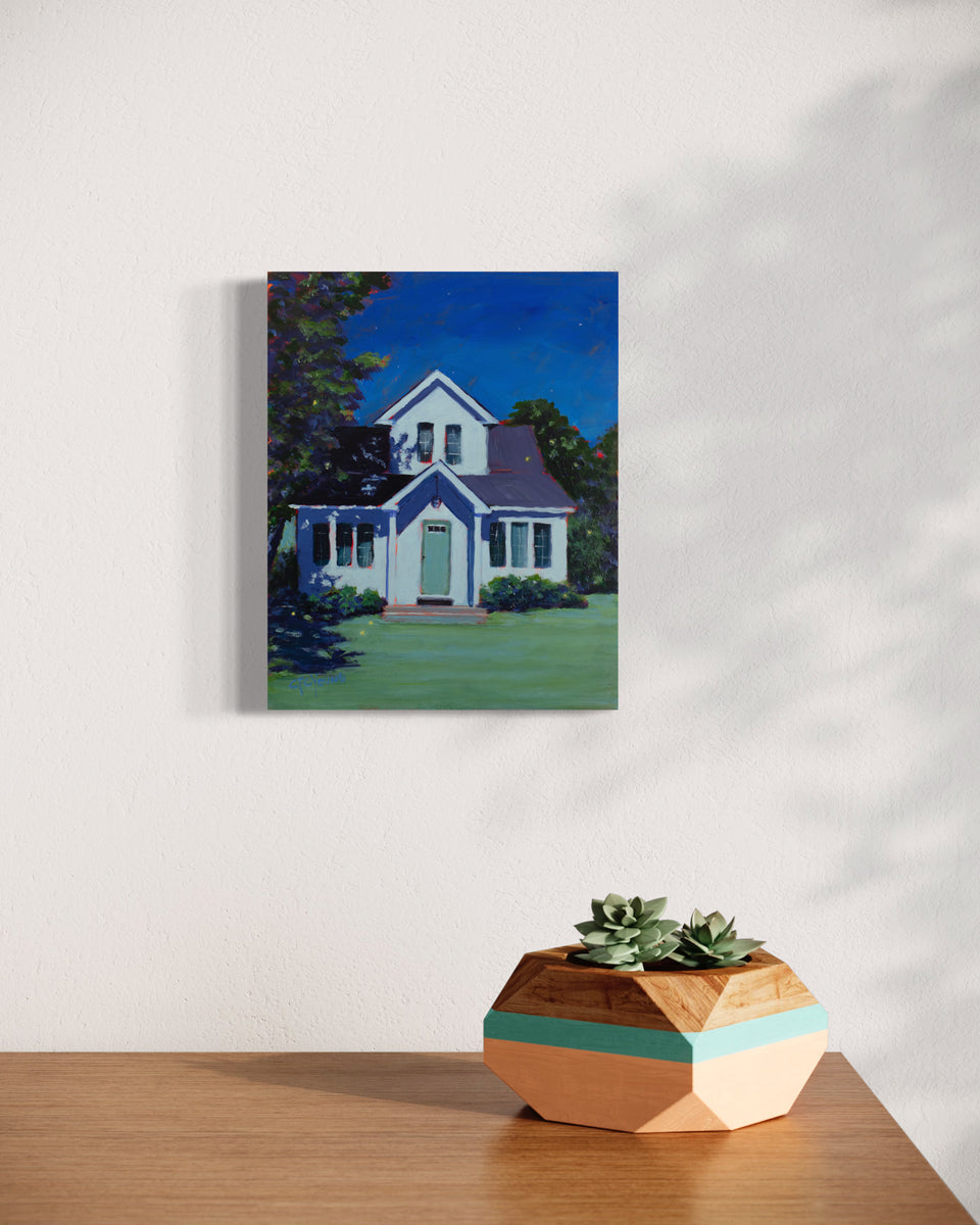 A landscape painting of a white house, lit by moonlight with a dark sky and a large tree to the left side by Carol Young hangs on a white wall above a wood surface with a potted succulent arrangement.