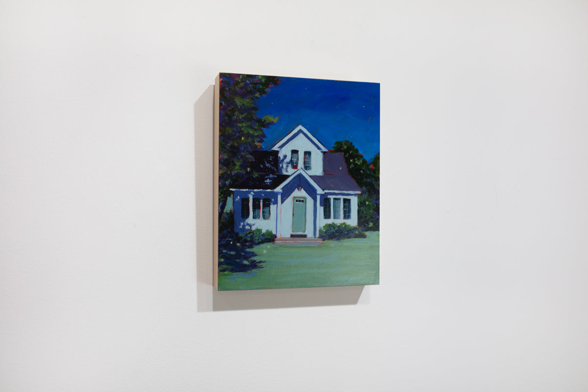 A left side view of a landscape painting of a white house, lit by moonlight with a dark sky and a large tree to the left side by Carol Young hangs on a white wall.