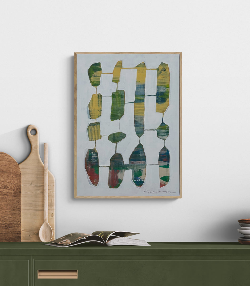 An abstract painting framed in a light wood frame hangs on a white wall above a green table with kitchen decor. 