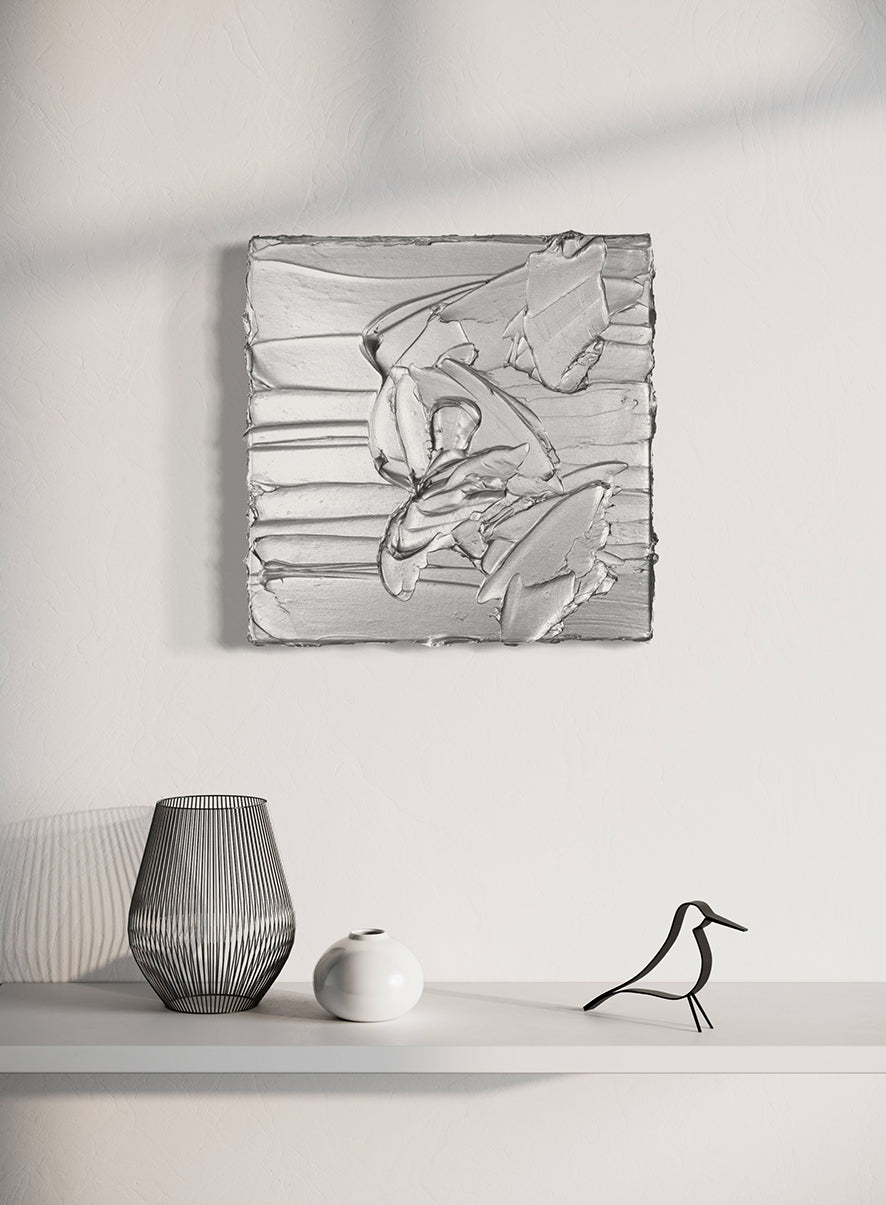 A silver metallic textured painting by Teodora Guererra hanging on a wall above a white shelf with decorative objects.
