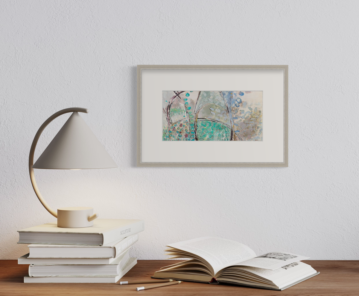 A teal, beige and mauve abstract landscape print in a silver frame with a mat hangs on a white wall above a natural wood surface with a stack of books holding a small lamp and an open book.