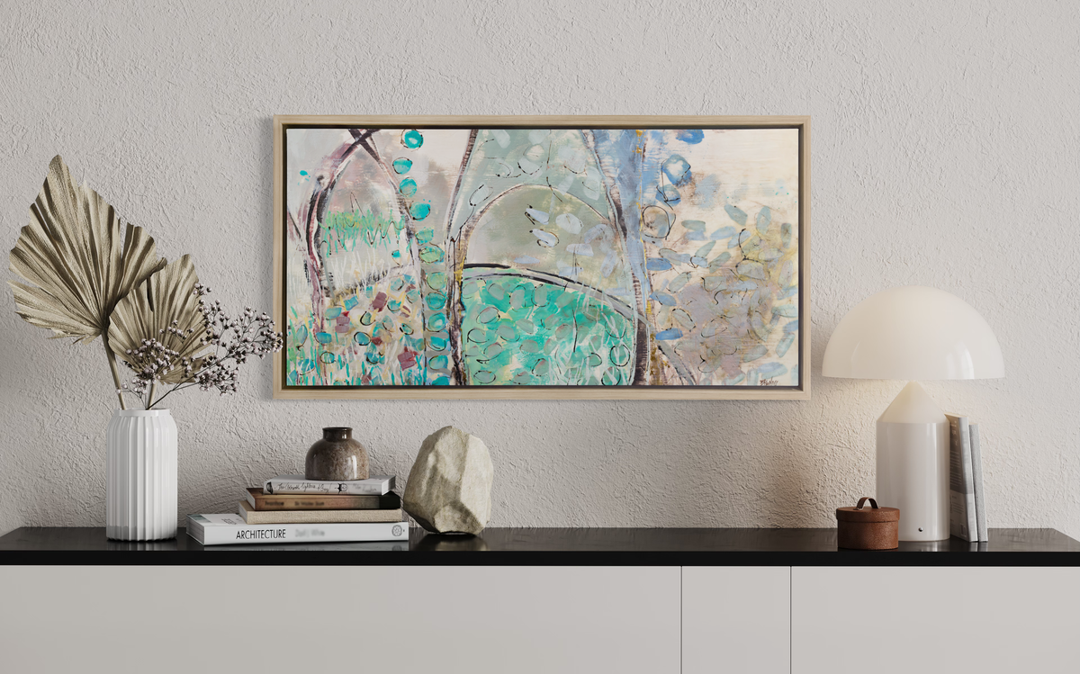 A teal, beige and mauve abstract landscape print in a white oak wood floater frame hangs on a white wall above a black surface with a white lamp, a stack of books, a rock and a white vase with decorative stems. 
