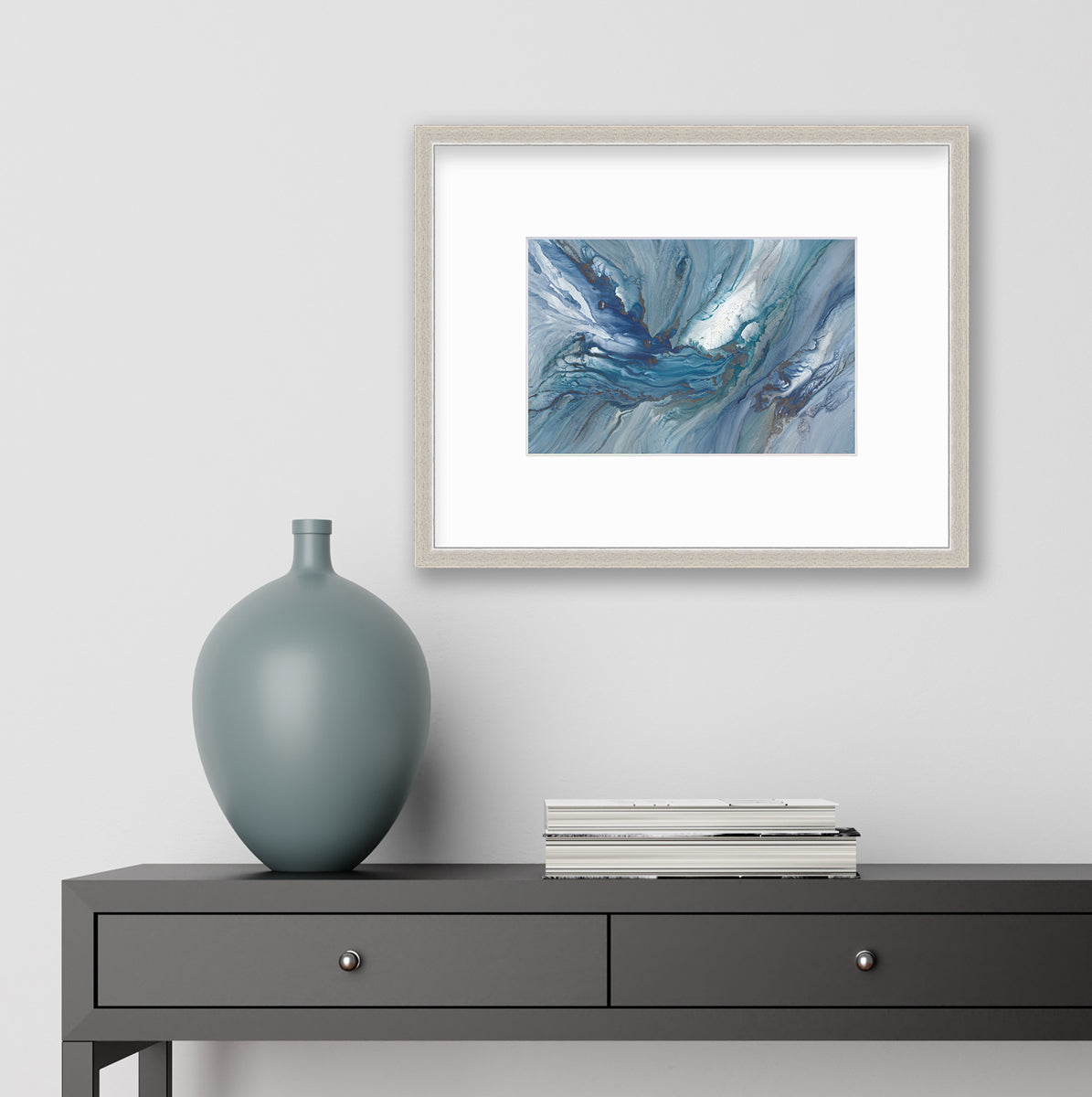 A blue, teal and white abstract print in a silver frame with a mat hangs on white wall above a black console table with a stack of books and a muted teal vase.