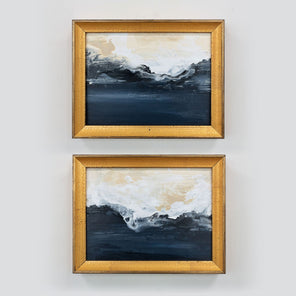 Two blue and white abstract paintings framed in gold frames hang on a light wall. 