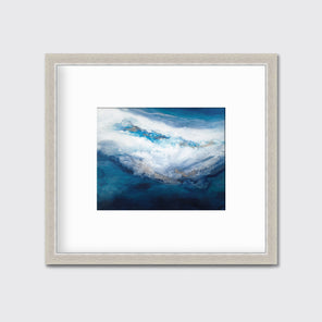 A blue, white, gold and teal abstract print in a silver frame with a mat hangs on a white wall.