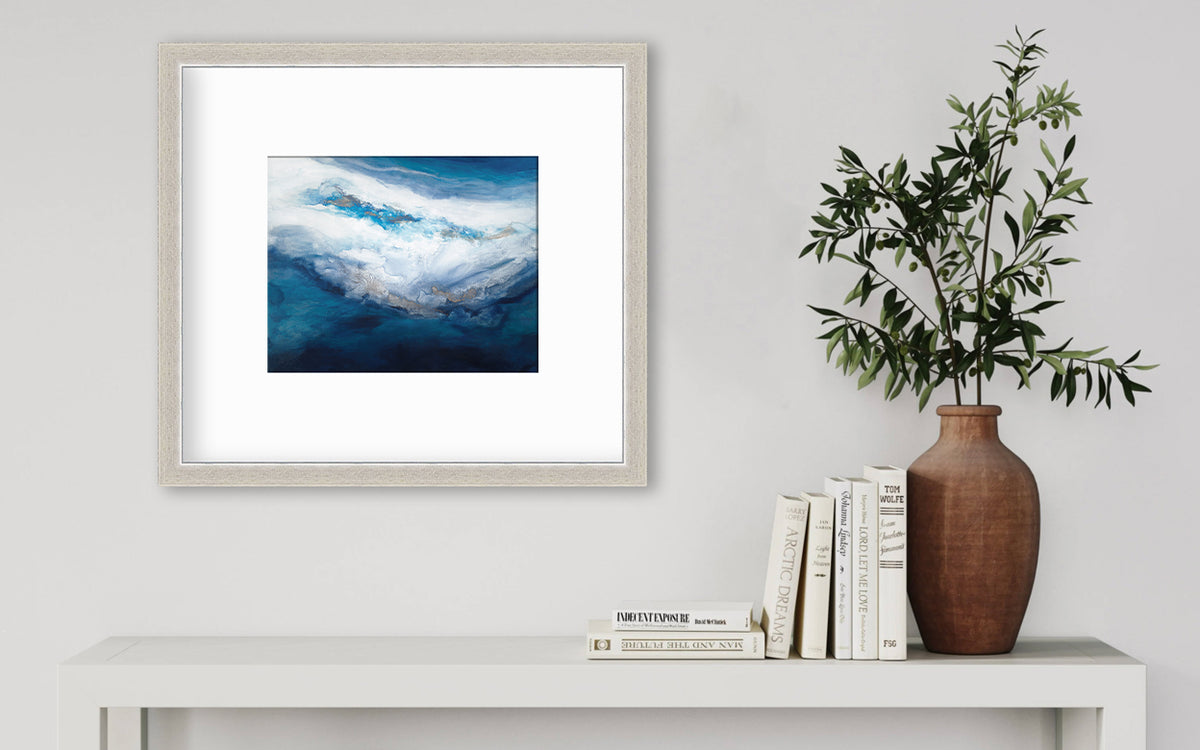 A blue, white, gold and teal abstract print in a silver frame with a mat hangs on a white wall above a white console table with a stack of books and a terracotta vase with greenery stems.