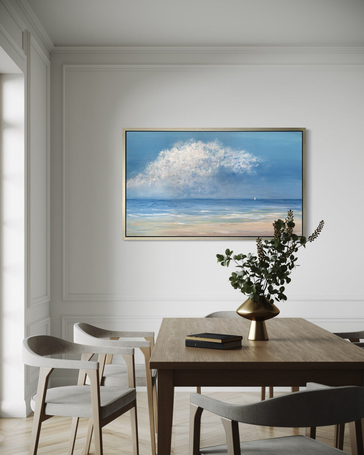 A painting of a contemporary seascape with one loan sailboat hangs on a wall in a warm silver floater frame above a dining table with a centerpiece of dried flowers. Chairs in white felt surround it and sunlight streams in from the left.