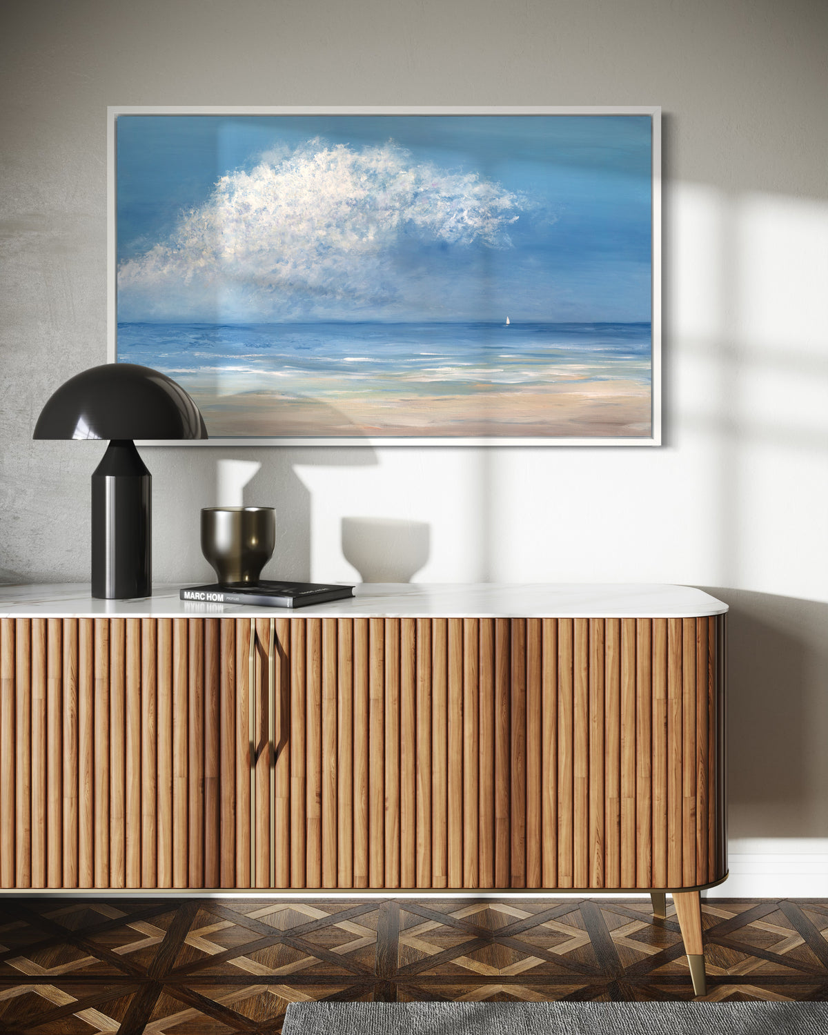 A painting of a contemporary seascape with one loan sailboat hangs on a wall in a white floater frame above a credenza with black lamp and accessories.