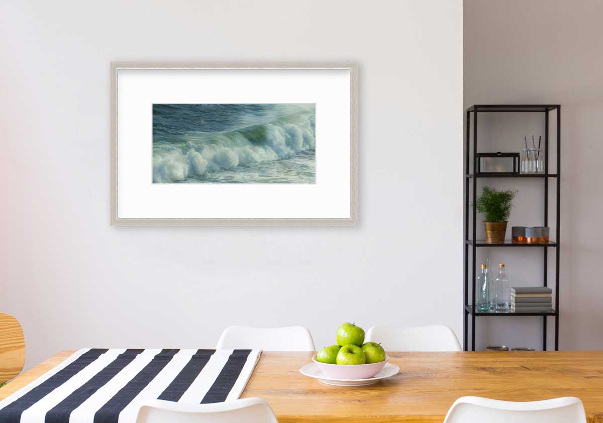 A blue, teal, white and light green abstract water wave print in a silver frame with a mat hangs on a white wall above a wood table with a blue and white striped table runner, a bowl of green apples and four white chairs.