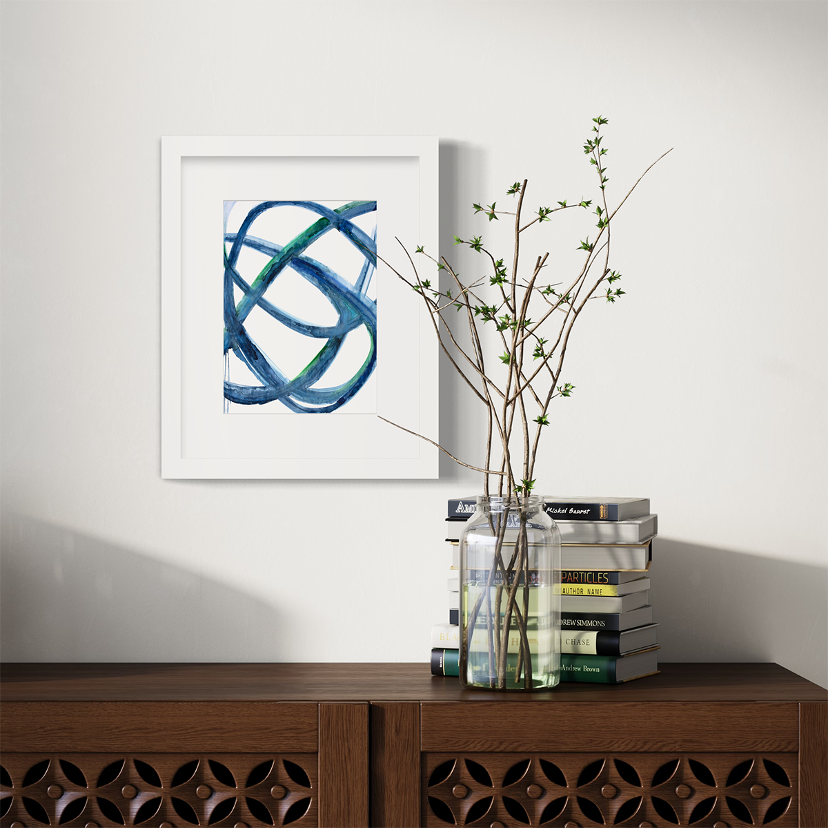 A blue, green and white abstract print in a white frame with a mat hangs on a white wall above a dark wood surface with a stack of books and a clear jar with greenery stems.