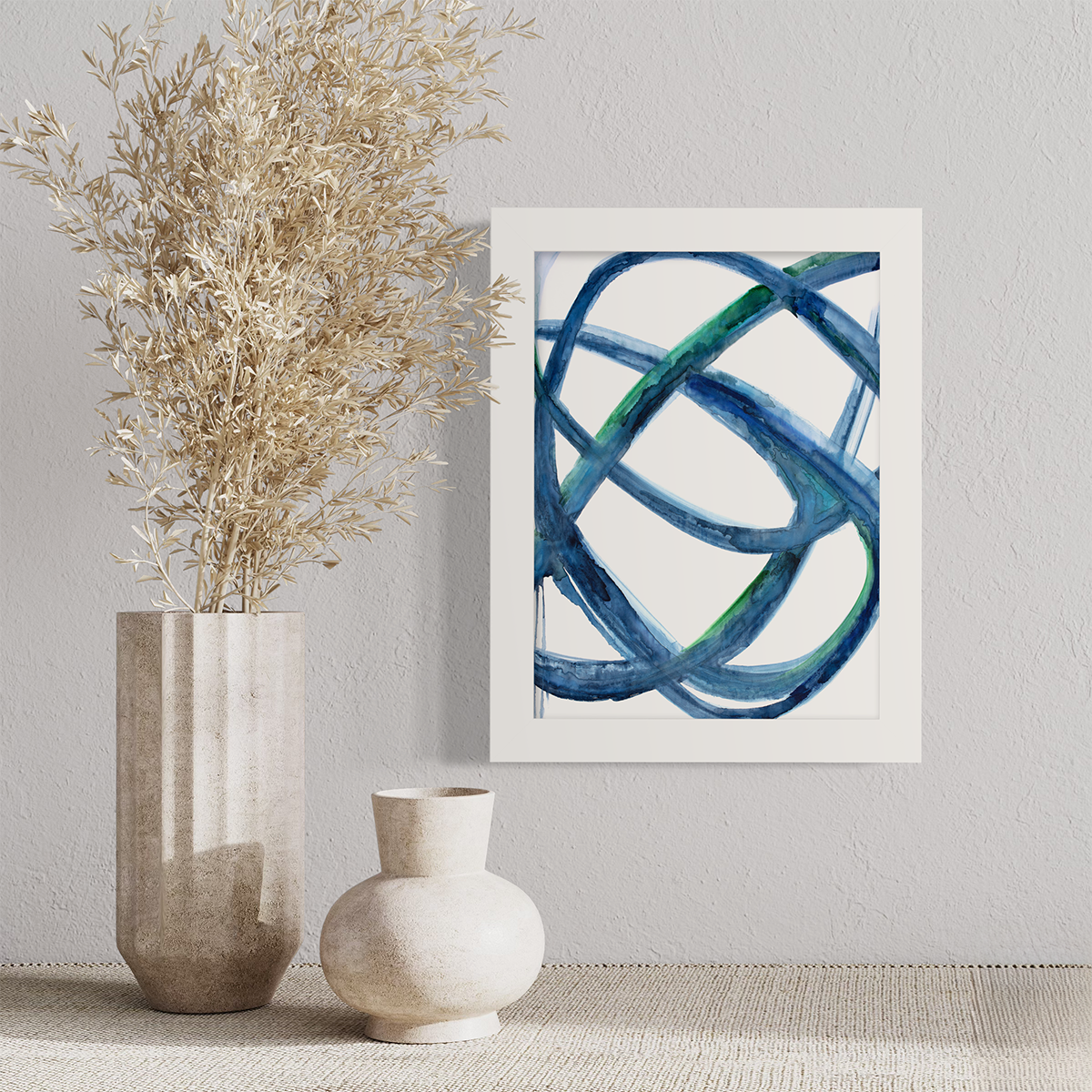 A blue, green and white abstract print in an unmatted white frame hangs on a white wall above a linen surface with a light beige vase with wheat stems and small beige vase.