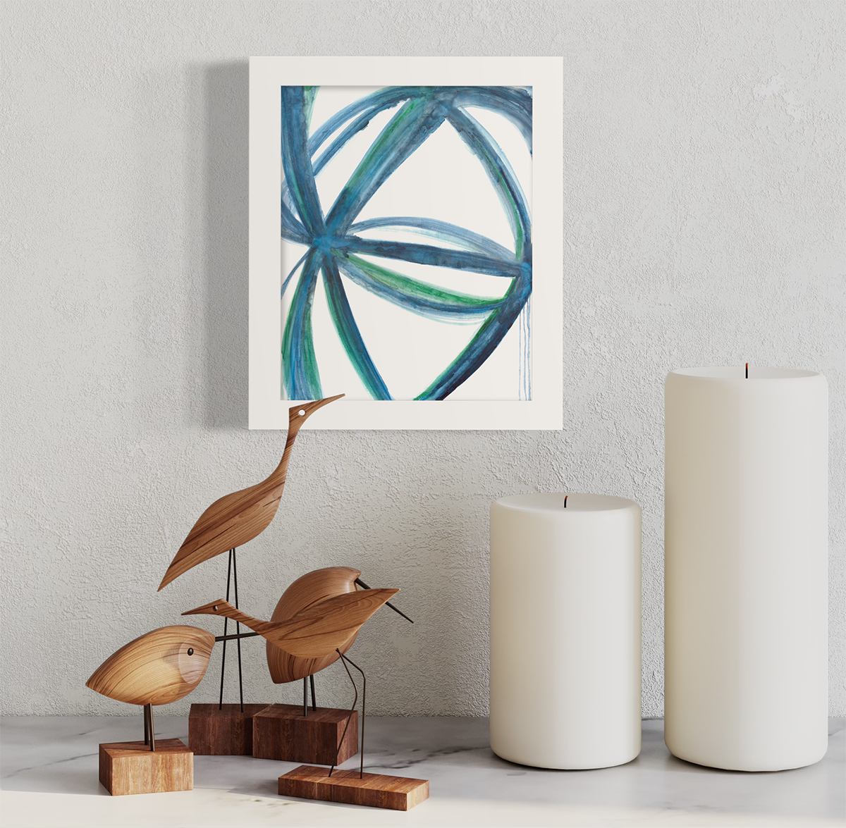 A blue, green and white abstract print in an unmatted white frame hangs on a white wall above white pillar candles and wooden bird figurines.