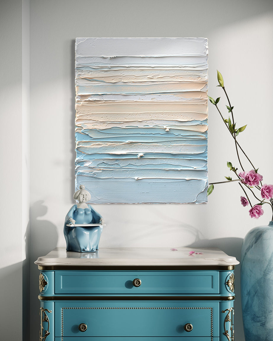 A thickly painted painting in white, orange sherbet and teal blue by Teodora Guererra hangs on a white wall over an ornate teal end table with white marble top. To the right is a marble vase with pink flowers and sitting on the table is a figurine of an asian woman in a teal dress. The painting is like a wall sculpture.