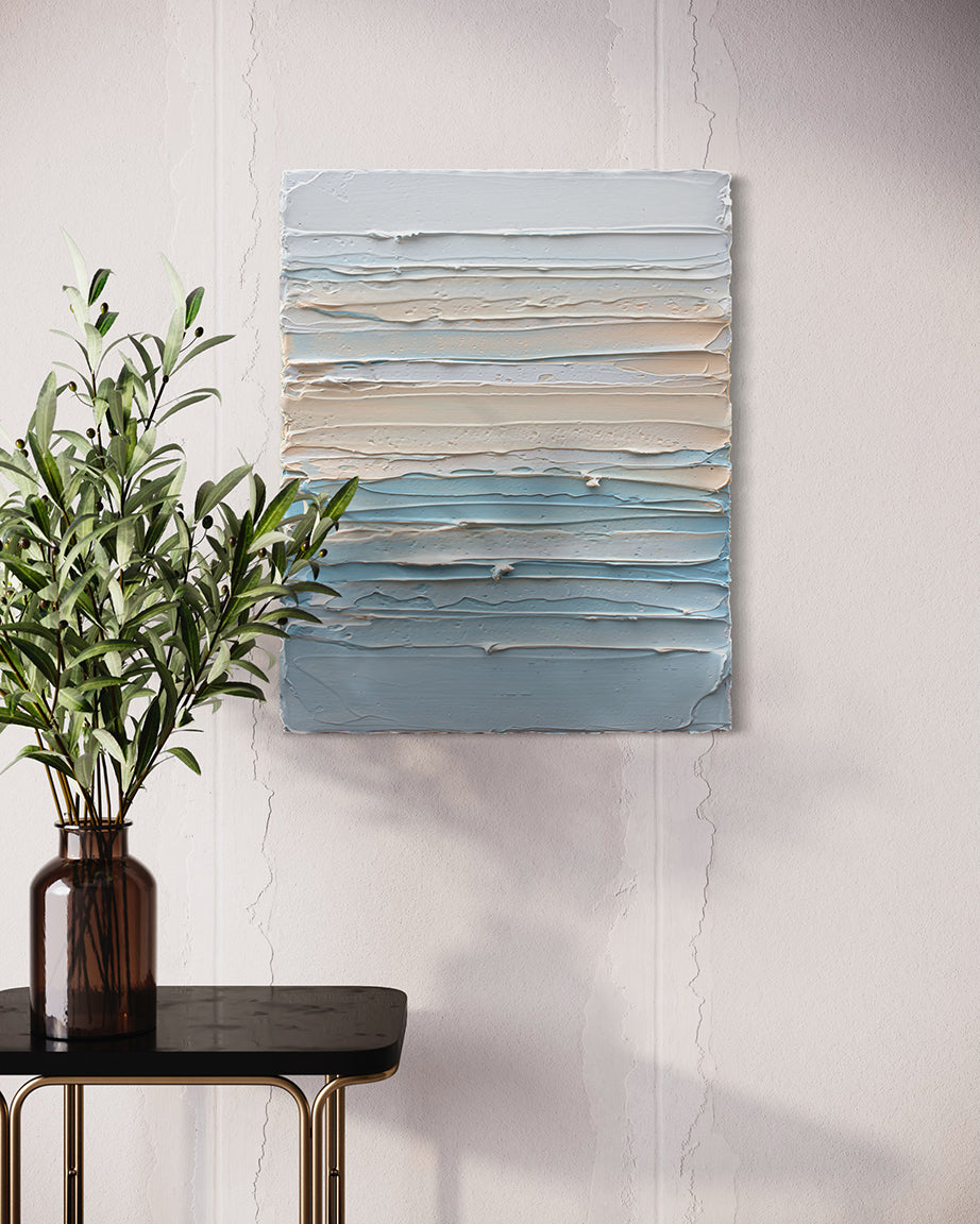A thickly painted painting in white, orange sherbet and teal blue by Teodora Guererra hangs on a white stucco wall over and to the left of a dark marble end table with brass legs. On the table is an amber glass vase with eucalyptus branches.
