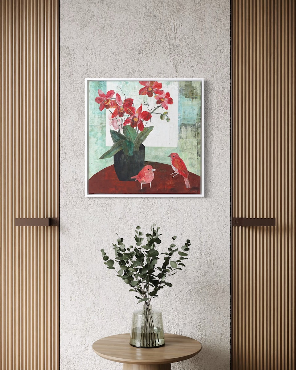 A framed collage painting depicting red flowers and birds hanging on a textured light gray wall, styled above a small round wooden table with a vase of greenery.