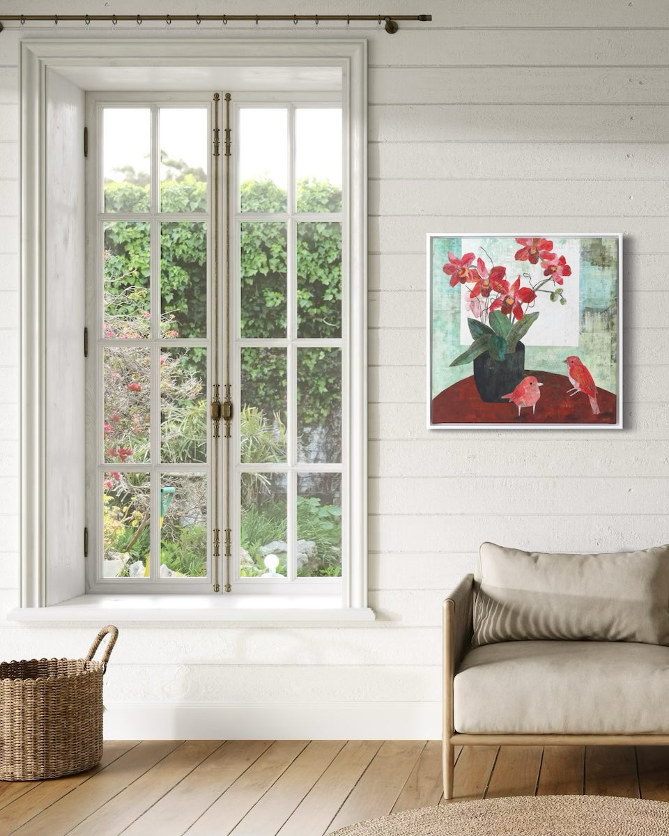 A framed collage painting depicting red flowers in a black pot and two red birds, hung on a white wood-paneled wall next to a large window in a bright, modern room with a beige armchair.