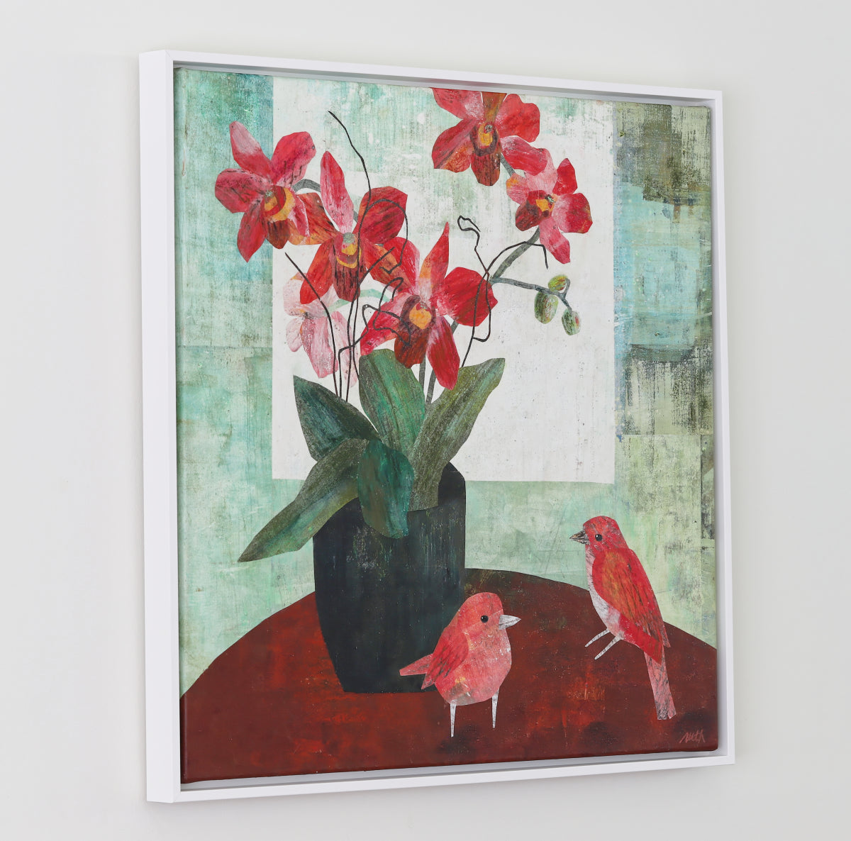 A framed square collage painting depicting red orchids in a black vase with two small red birds on a dark red surface, set against a textured light green and white background, hung on a light gray wall.