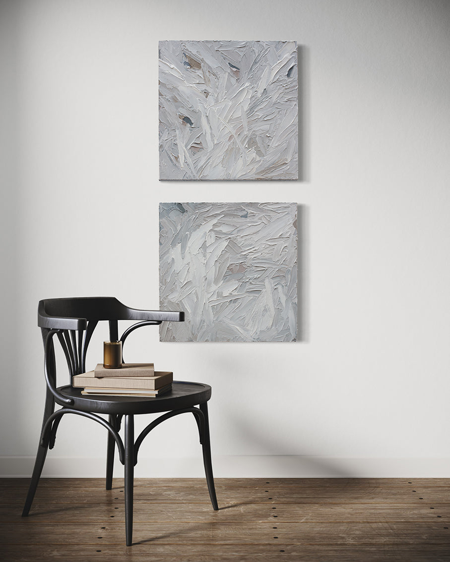A pair of thickly textured paintings in grey, white, blue grey and plum grey by Teodora Guererra hanging on a white wall in front of a brown chair with books and a candle sitting on it.