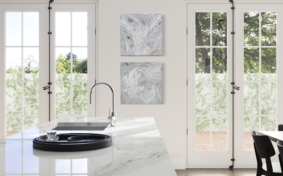 A pair of thickly textured paintings in grey, white, blue grey and plum grey by Teodora Guererra hang on a white wall in a kitchen. There is a white marble island in front of the paintings on the wall and on either side are french doors leading out to the back yard.