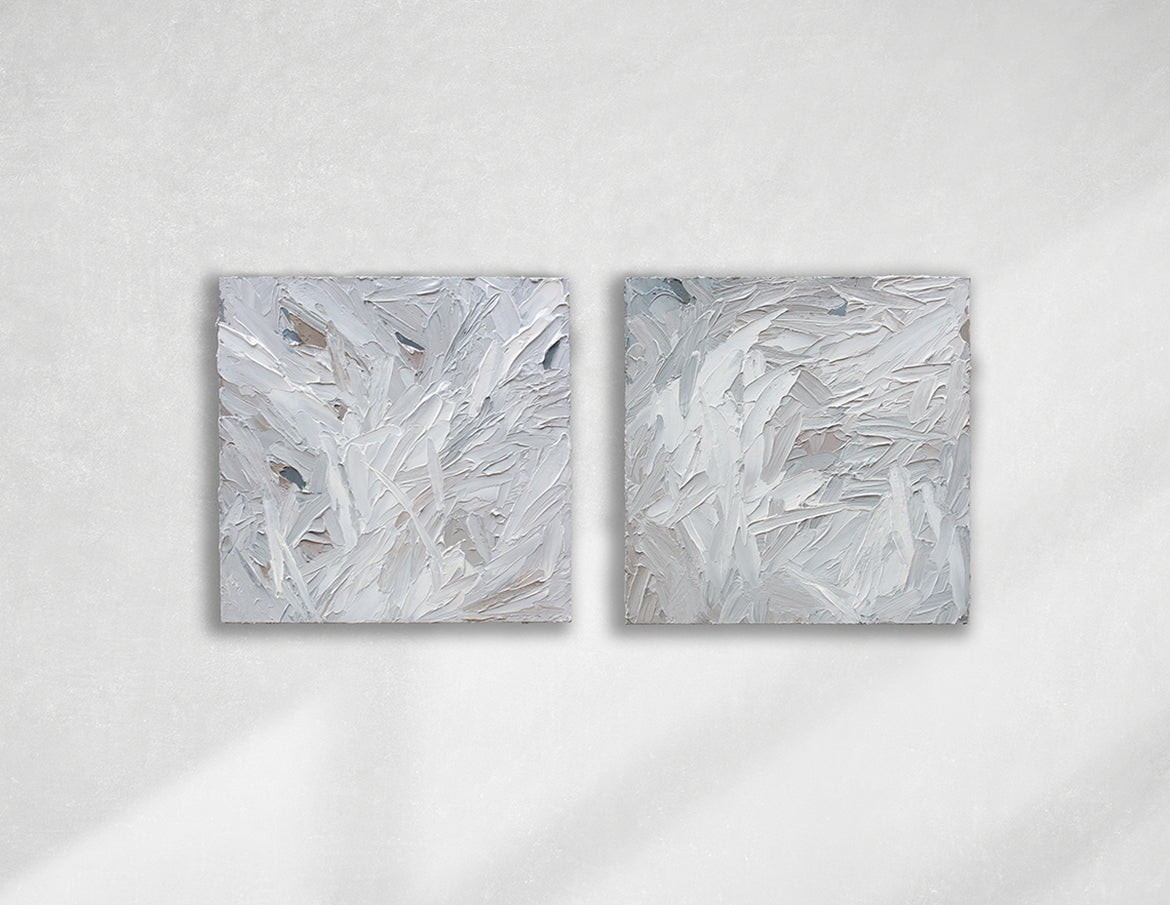 A pair of thickly textured paintings in grey, white, blue grey and plum grey by Teodora Guererra hanging on a white wall.
