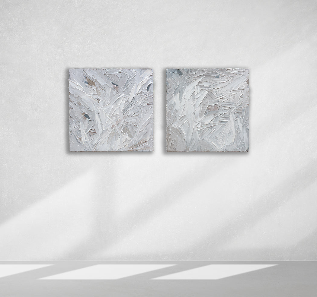 A pair of thickly textured paintings in grey, white, blue grey and plum grey by Teodora Guererra hanging on a white wall.