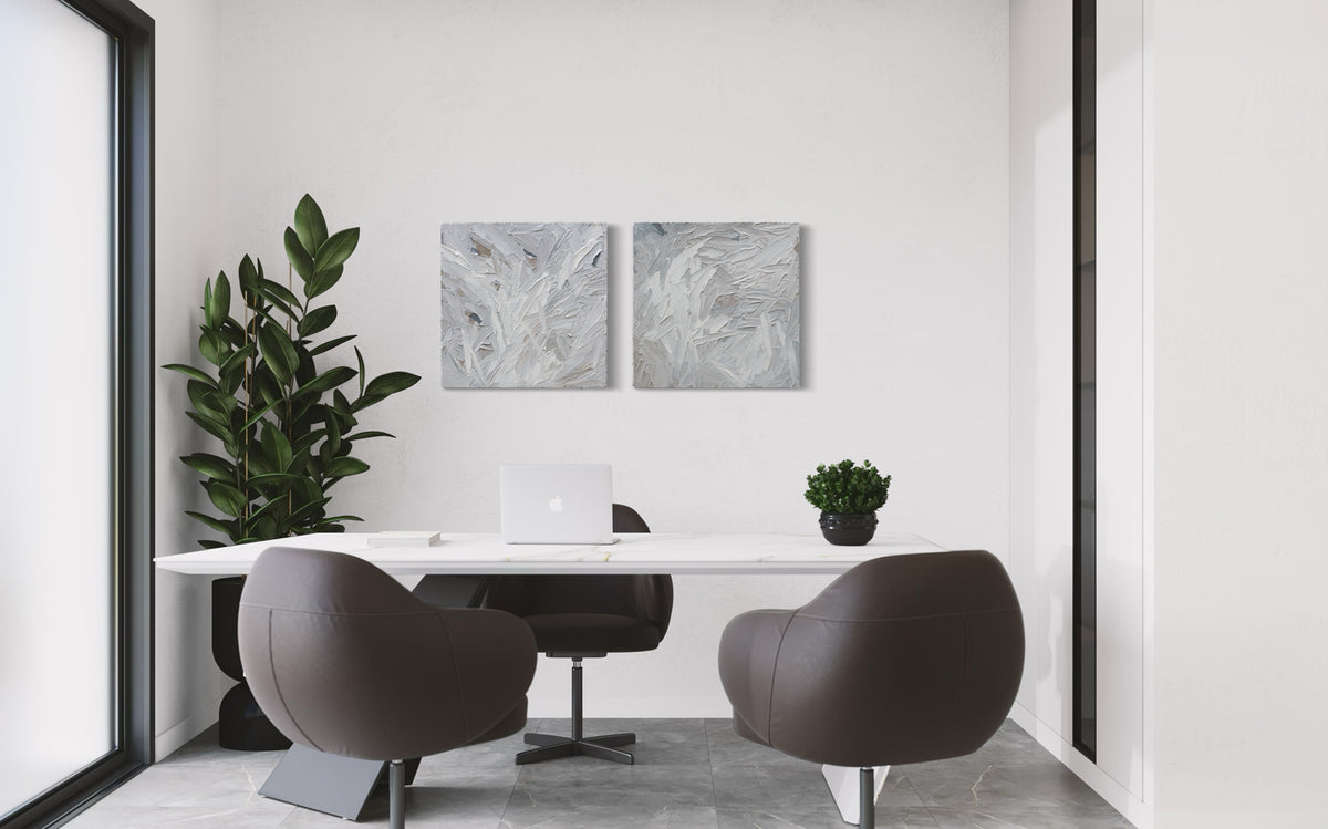 A pair of thickly textured paintings in grey, white, blue grey and plum grey by Teodora Guererra hanging on a white wall behind a desk.
