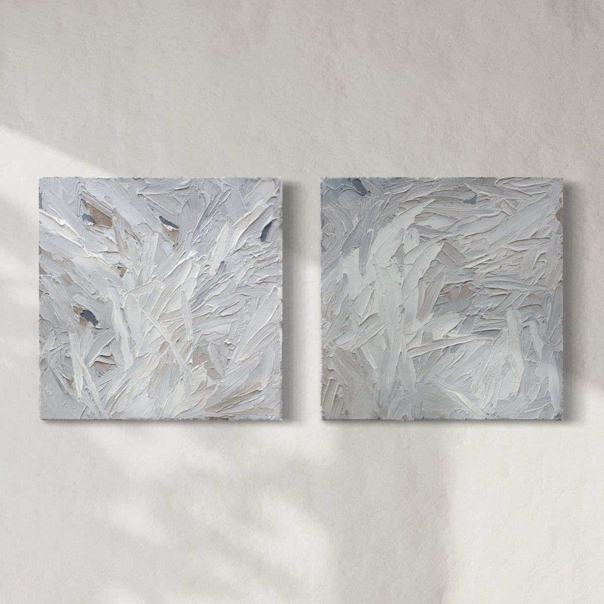 A pair of thickly textured paintings in grey, white, blue grey and plum grey by Teodora Guererra hanging on a white wall.