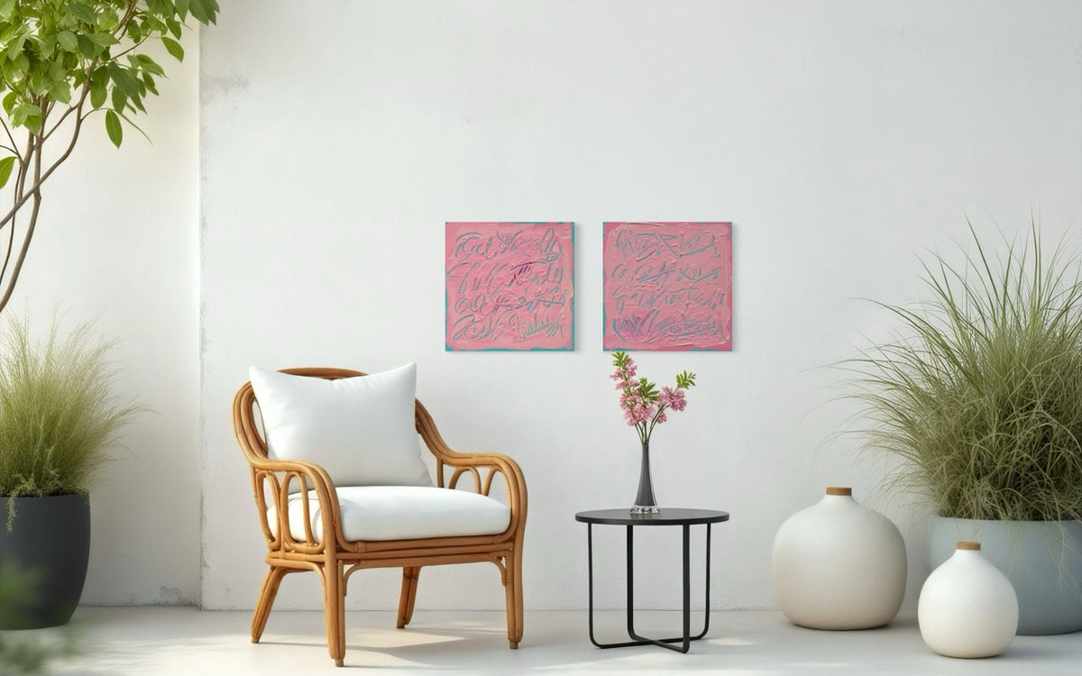 Two square pink text based paintings with writing into the surface with various tools, carving fluid script that reveals the teal color hidden below. Displayed side-by-side on a white wall in a room with wicker chair and table with flowers.